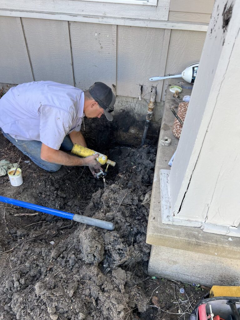 A plumber repairing a water line in an excavation beside a house